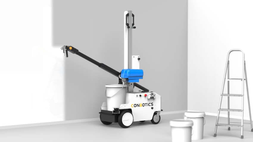 An autonomous painting robot stands in an empty interior space in front of a wall. The robot is mounted on wheels and equipped with an extendable arm that applies paint to the wall. Paint buckets and a ladder are placed nearby. The scene illustrates the use of robotics in professional painting work.
