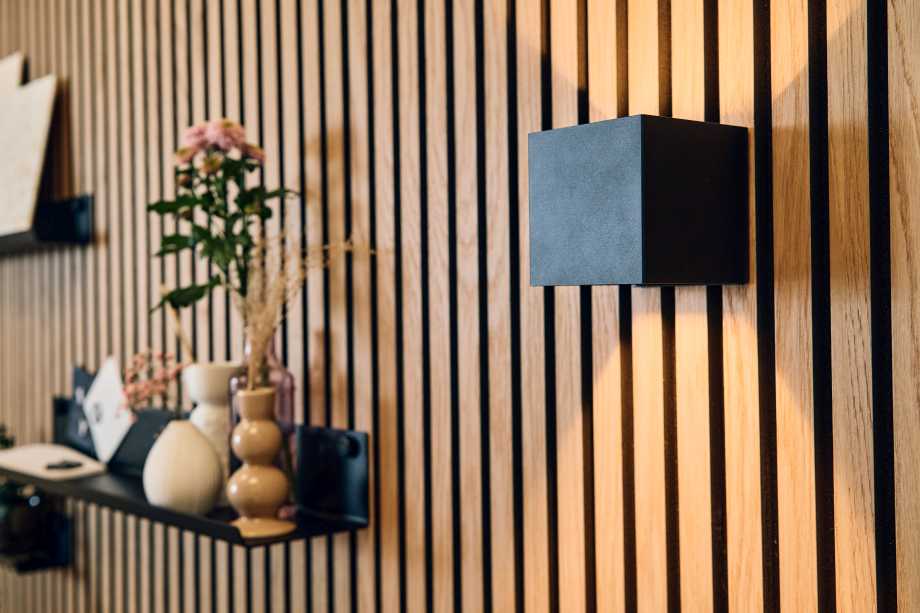 Moderne Holzpaneel-Wand mit vertikalen schwarzen Lamellen. An der Wand ist ein minimalistischer schwarzer Würfel-Wandleuchte montiert, die nach oben und unten Licht abstrahlt. Im Hintergrund befinden sich dekorative Vasen mit Trockenblumen und eine kleine Blumenvase mit rosa Blüten auf einem schwarzen Wandregal. Produktneuheit des Ausstellers ter Hürne auf der DOMOTEX 2026 in Hannover