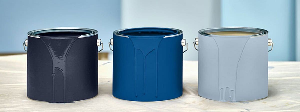 Three open paint cans are placed side by side on a light surface. The colors shown are dark blue, rich blue-green, and light gray. The image visualizes contemporary color trends and design shades.