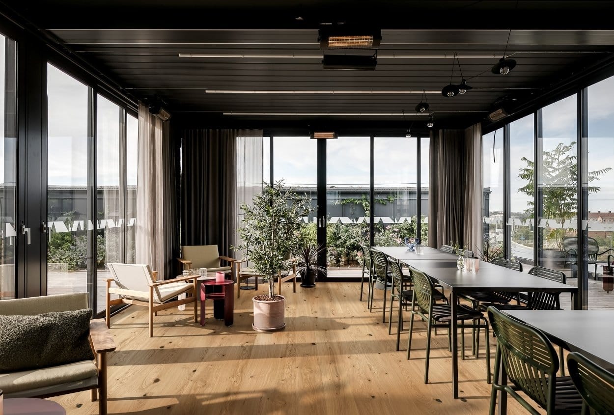 View into a modern interior space with large glass fronts and a natural wood floor. The interior showcases a residential, sustainable design approach with light materials and clean furnishings.